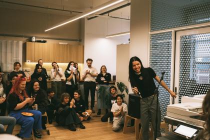 A group of people are in an office space. One person is holding a microphone up front and smiling at someone out of frame. Some others in the crowd are clapping. Others are talking to each other.