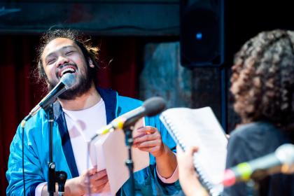 A man wearing a bright blue jacket is smiling while singing into a microphone. Another person is facing them we can see the back of their curly hair. They are both holding up notebooks as though this is a rehearsal.