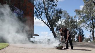 A first nations man is doing a smoking ceremony in an outdoor space with gumtrees in the background. 