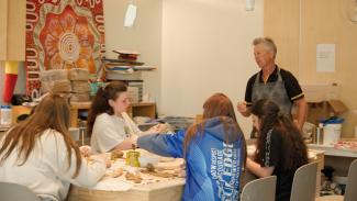 Four kids are sitting around a table in an art room. The teacher is overseeing their clay making experience. 