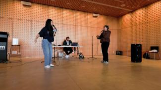 Three people are standing in sound proof room. One is playing keyboard, the other two are singing into microphones while look at each other.