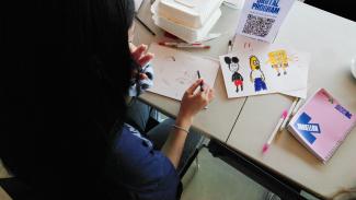 A birds eye shot of a person sketching a face. The piece of paper next to them has a crayon drawing of Mickey Mouse, Homer Simpson and Spongebob Squarepants. 