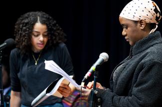 Front of frame is a woman wearing a bandanna is looking at a piece of paper and standing next to a microphone.. Behind her is another woman doing the same thing. 