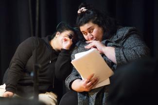 Two people are huddled behind a script whispering cheekily to each other. 