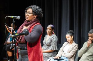 A woman wearing a long bright red vest, maroon scarf and hoop earrings is standing in front of a microphone reading from her phone. Behind her sits a row of people all looking at her or at pieces of paper. 