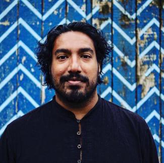 South Asian consultant, Shankar Kasynathan. Shankar is standing in front of a blue patterned wall smiling at the camera