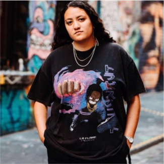 Lyle Makepeace, a young Pasefika consultant, is standing in front of a wall of colourful grafiti facing the camera with their hands in their pockets. Lyle is wearing a silver chain necklace and black t-shirt and pants.
