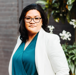 Pasefika consulant, Rita Seumanutafa, is standing in front of a brick wall earing a teal top and white cardigan smiling at the camera. There are some white flowers behind her right shoulder.