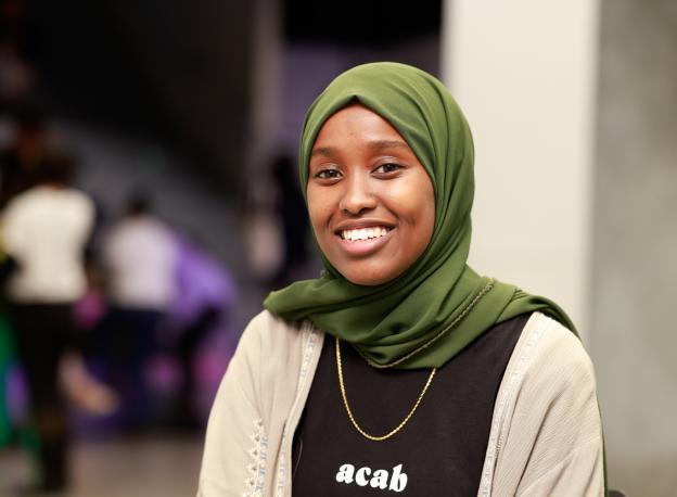 Portrait shot of Idil Ali smiling to camera. They are wearing a gold necklace, a top with acab and a green hijab 