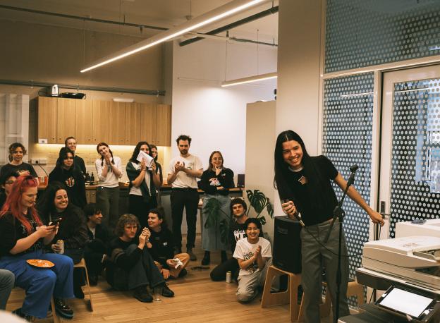 A group of people are in an office space. One person is holding a microphone up front and smiling at someone out of frame. Some others in the crowd are clapping. Others are talking to each other. 