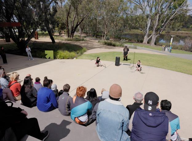 Camera is behind a crowd of people sitting on some steps watching a first nations ceremonial dance. 